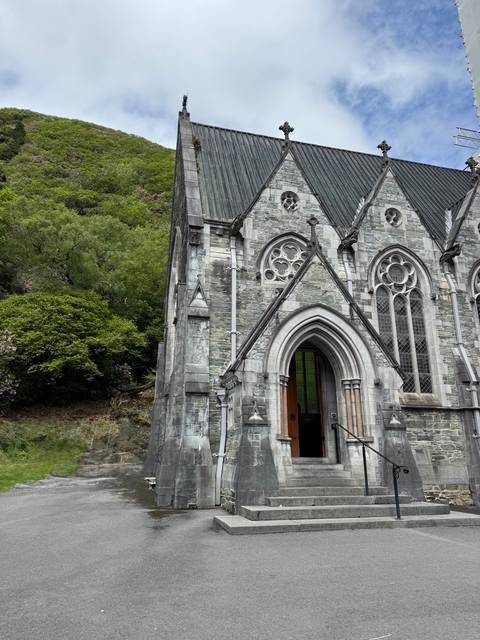 Partial view of a grey-stone neo-Gothic church façade surrounded by lush greenery.