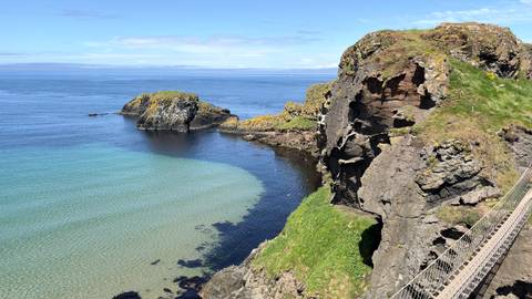 Clear turquoise cove beside rugged cliffs and a rope bridge viewpoint on a sunny day