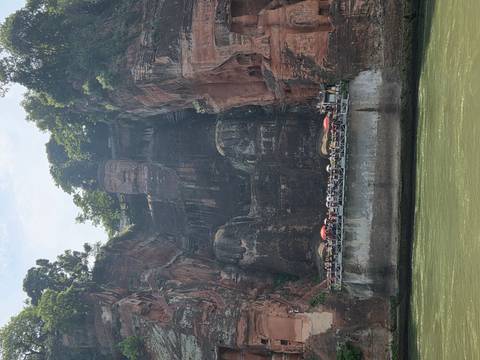 Massive seated stone Buddha carved into a red cliff towering above a river with tourists on a viewing platform.