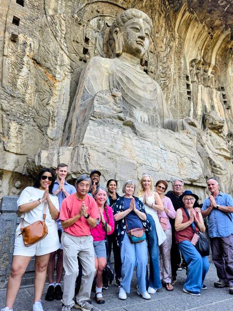 Tour group poses with prayer hands in front of a large ancient stone carving of a Buddha figure.