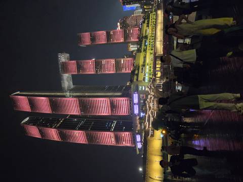 Nighttime city promenade with crowds beneath futuristic pink-lit skyscrapers connected by a skybridge.