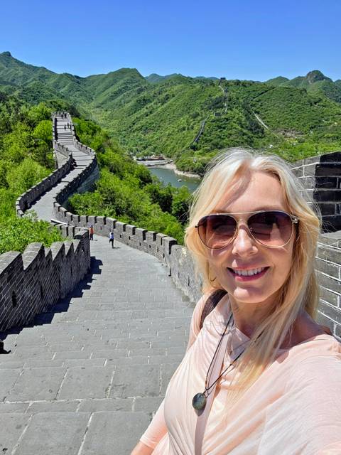 Woman takes a selfie on the battlements of the Great Wall overlooking green hills and a river bend.