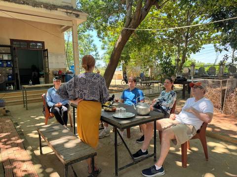 Small group listening to a host while seated outdoors under trees with tea set on a wooden table.