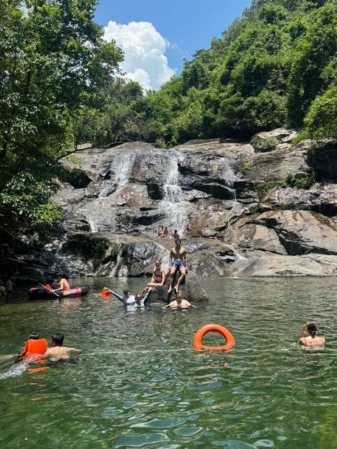Travelers swimming and posing on rocks at a jungle waterfall pool with kayaks nearby.