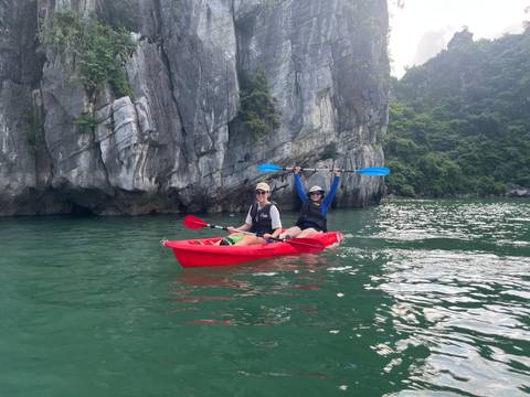 Two travelers in a red kayak celebrating with paddles raised amid green waters and towering limestone cliffs.