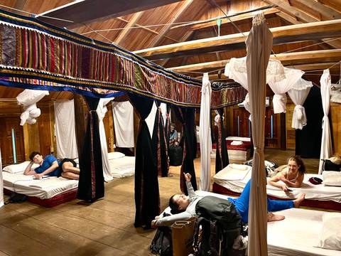 Rustic wooden dorm with travelers lounging on beds separated by colorful fabric drapes and mosquito nets.