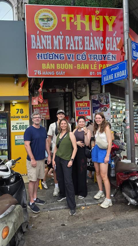 Family standing on a busy Hanoi street lined with small shops, signs, and scooter parts.