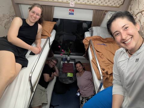Four travelers smiling inside an overnight train cabin with bunk beds and a small table.
