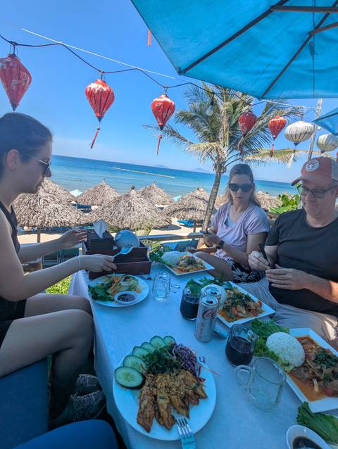 Family enjoying Vietnamese dishes at a beachfront restaurant under palm trees and thatched umbrellas.
