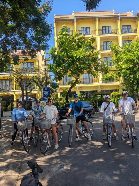 Six cyclists posing with bikes on a sunny street lined with bright yellow buildings in Hoi An.