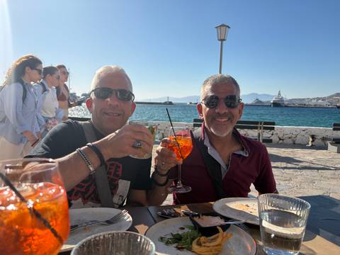 Two men raise colourful cocktails at a sunny seaside café with yachts and blue water behind them.