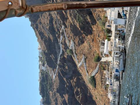 Steep zig-zagging path climbs a rocky cliffside above whitewashed buildings on the Aegean coast.