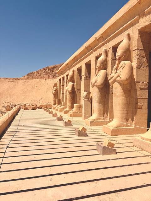 Row of monumental sandstone statues guarding a terrace against a backdrop of desert cliffs and blue sky.