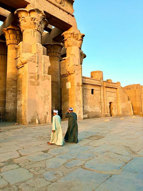 Two local men walk past towering carved temple walls bathed in soft evening light.