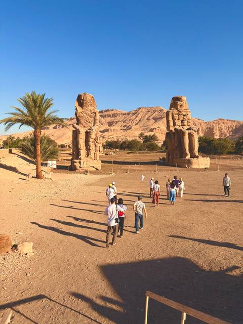 Visitors gather before the massive Colossi of Memnon with rugged desert mountains in the background.