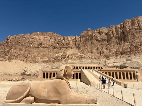 A sphinx statue overlooks tourists ascending the grand staircase of Hatshepsut Temple set beneath towering cliffs.