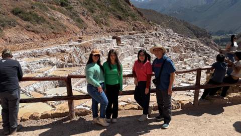 Four travellers pose on a viewpoint overlooking the terraced salt ponds of Maras set against an Andean mountain backdrop.