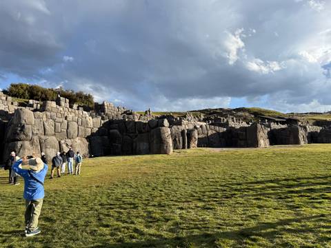 Wide lawn fronts the massive stone walls of the Sacsayhuaman fortress as tourists explore under dramatic clouds.