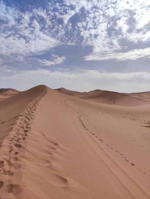 Sharp ridge of a sand dune lined with footprints under a patchy blue sky.