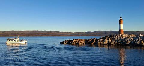 Red-and-white lighthouse on rocky islet with tourist boat on calm blue channel at golden hour.