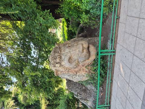 Weathered stone sculpture of a lion’s head partially surrounded by greenery in a town park.