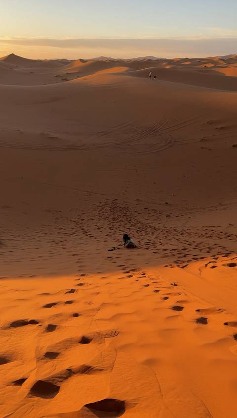Lone traveler sits on the vast slope of a desert dune at dusk, footprints dotting the sand.