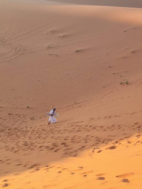 Local guide carries a sandboard up a steep dune, footprints trailing behind on the orange slope.