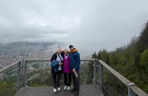 Three travelers pose on a viewing platform high above a foggy cityscape surrounded by mountains and water.