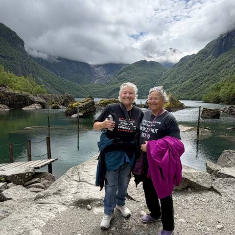 Two women smile in front of a serene fjord with steep green mountains and calm water.