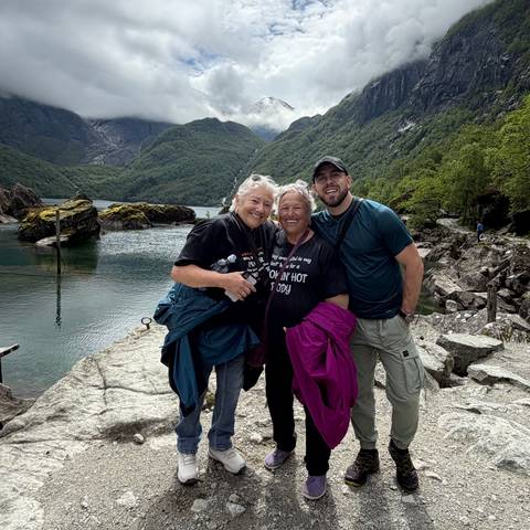 Three friends posing on rocky shore with green mountains and fjord waters in the background.