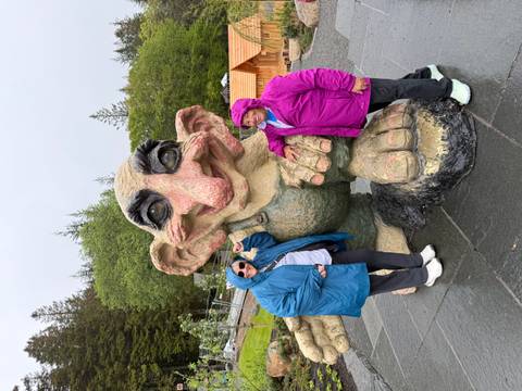 Two women pose with a large whimsical troll statue on a wet stone plaza.