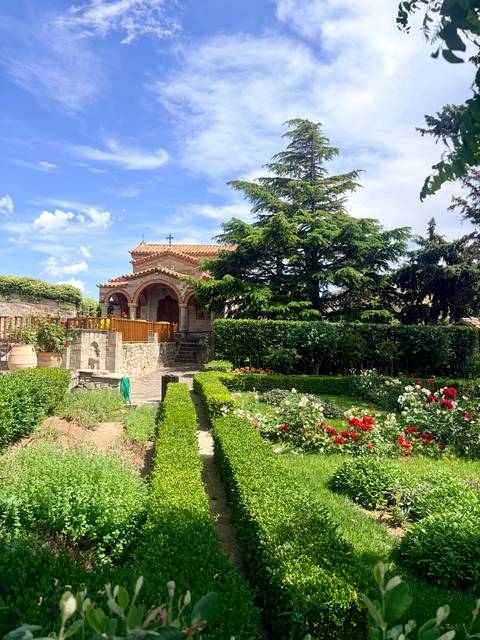 A small stone chapel sits amid manicured gardens with vibrant flowers and tall evergreens.