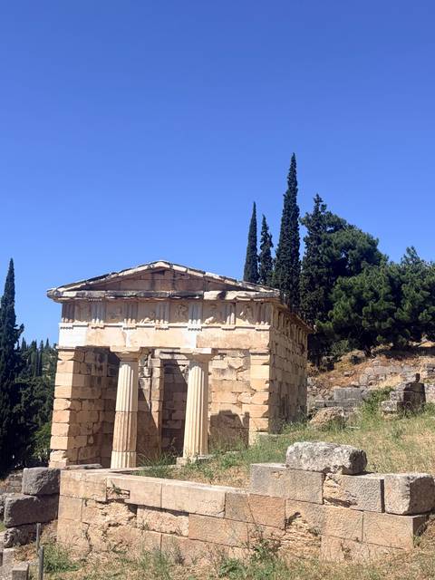 Ancient stone treasury with Doric columns stands beneath a clear blue sky surrounded by cypress trees.