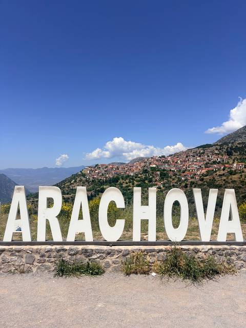 Large white letters spelling ARACHOVA overlook a hillside village and distant mountains under blue skies.