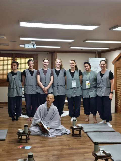 Temple-stay participants line up in grey vests next to a seated Buddhist monk.