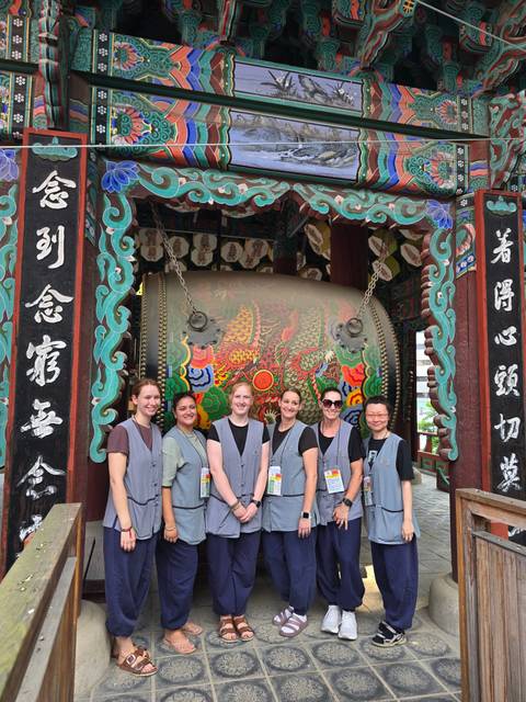 Group in temple uniforms pose beside a large ornate Korean temple drum.