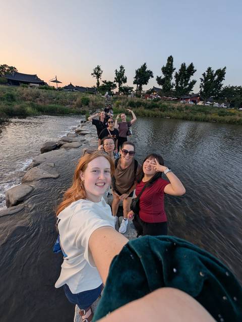 Friends laugh while crossing a river on stepping stones at dusk.