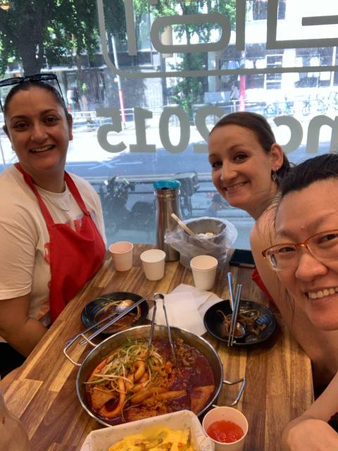 Three friends in aprons smile while enjoying noodle bowls at a casual eatery.