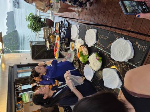 Cooking demonstration on a marble counter aboard a cruise boat, staff preparing fresh Vietnamese spring rolls with Halong Bay scenery in background.