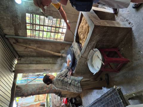 Local woman steams rice paper over a pot in a rustic workshop while another person tends the fire and rice husks.