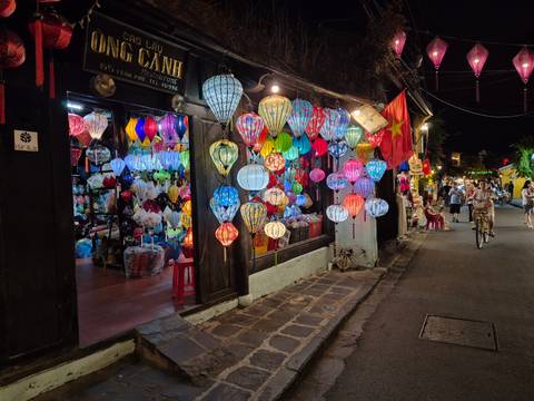 Night street in Hoi An decorated with dozens of vibrant silk lanterns outside a shop while pedestrians stroll by.