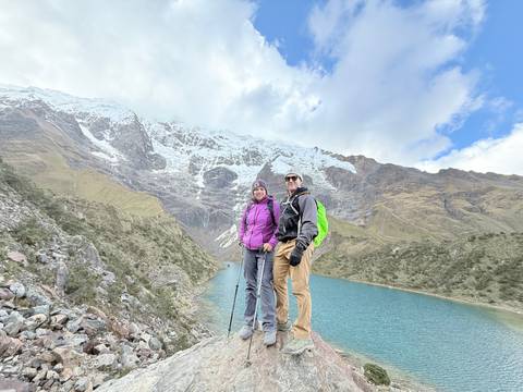 Couple stands beside turquoise Humantay Lake with snow-covered Andes peaks looming behind.