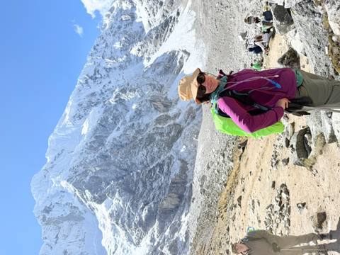 Female trekker smiles in front of a towering snow-capped Andean peak on a sunny day.