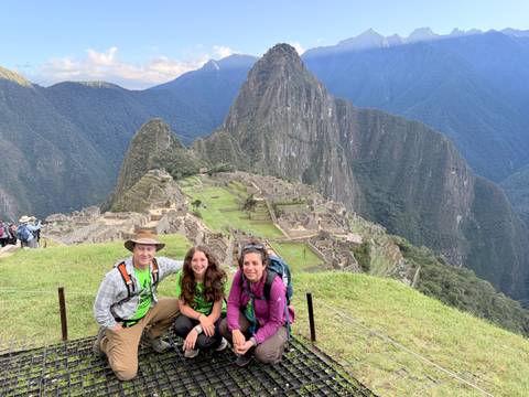 Three trekkers sit at the classic Machu Picchu viewpoint with the citadel and Huayna Picchu behind.