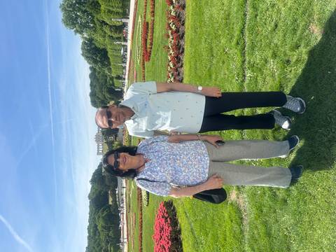 Couple stands in colorful flower gardens with the Gloriette of Schönbrunn Palace faintly visible in the distance.