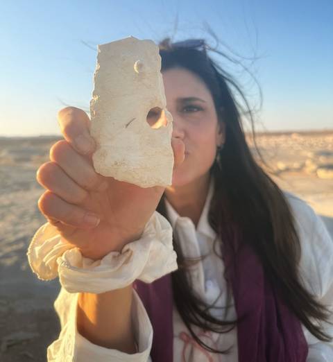 Woman holding a pale desert rock fragment toward the camera with arid landscape behind