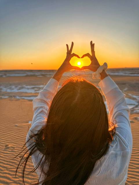 Back-lit woman forming a heart shape with her hands around the setting sun over desert flats
