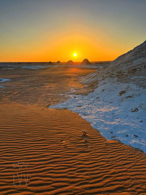 Rippled orange sand meeting pale chalk ridges with the sun setting behind distant formations