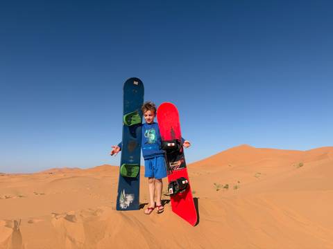 Child holding sandboards on a vast desert landscape under clear blue sky