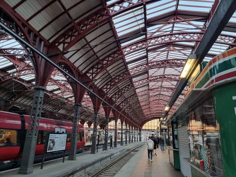 Interior of a historic iron-roofed railway station with red girders and waiting trains.
